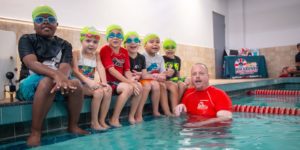 Children at swim class sitting along edge of pool with swim instructor in water