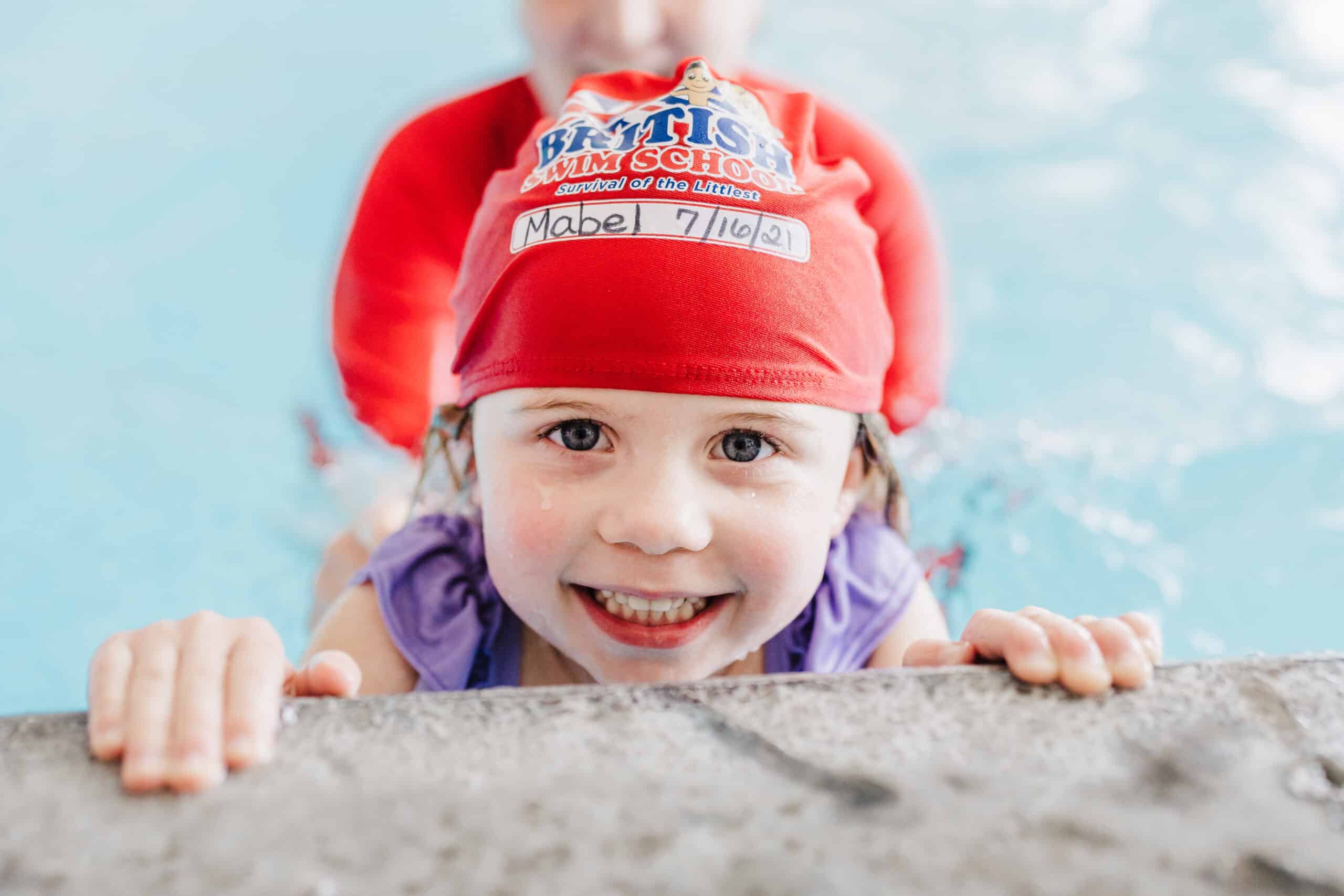 A smiling young girl in a red swim cap holds onto the pool edge during a swim lesson.