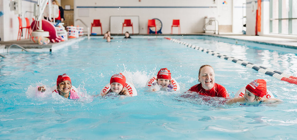 Four smiling children and an instructor swim happily in an indoor pool.