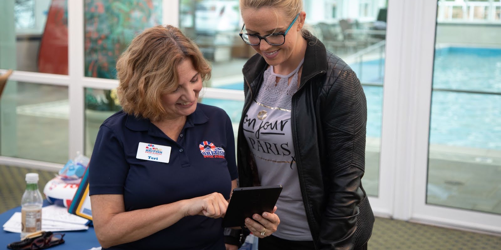A swim instructor helps a woman register on a tablet near an indoor pool.