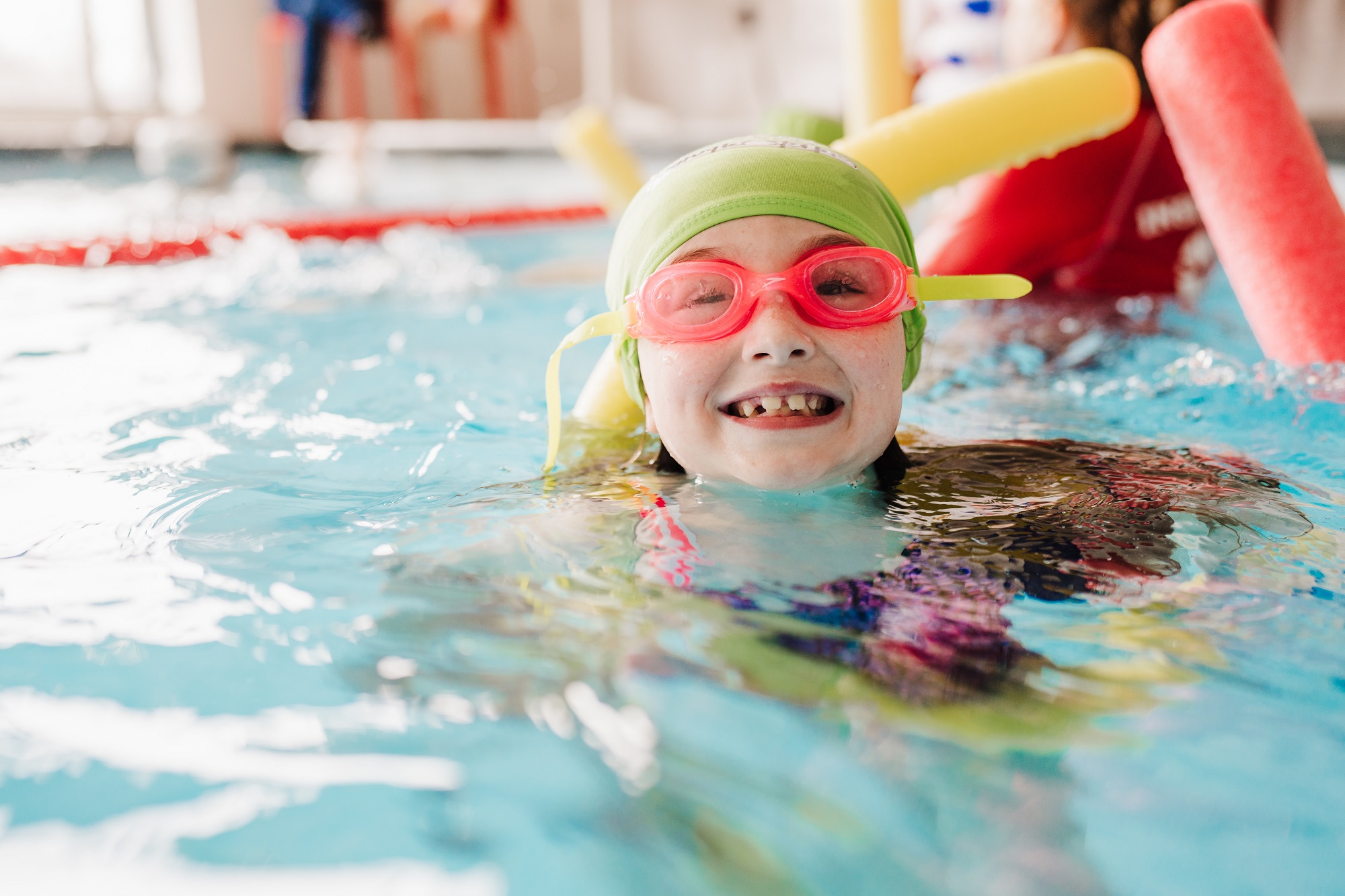 A happy child in a green swim cap and pink goggles smiles with missing teeth in a