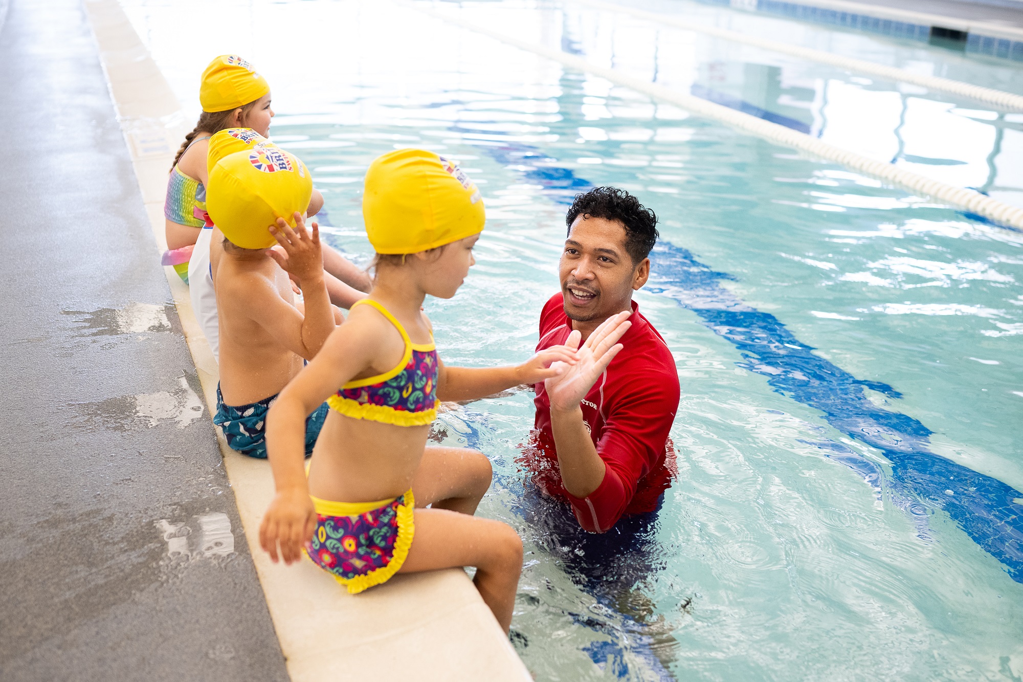 Smiling swim instructor in the pool demonstrates to young children in yellow caps sitting on the edge